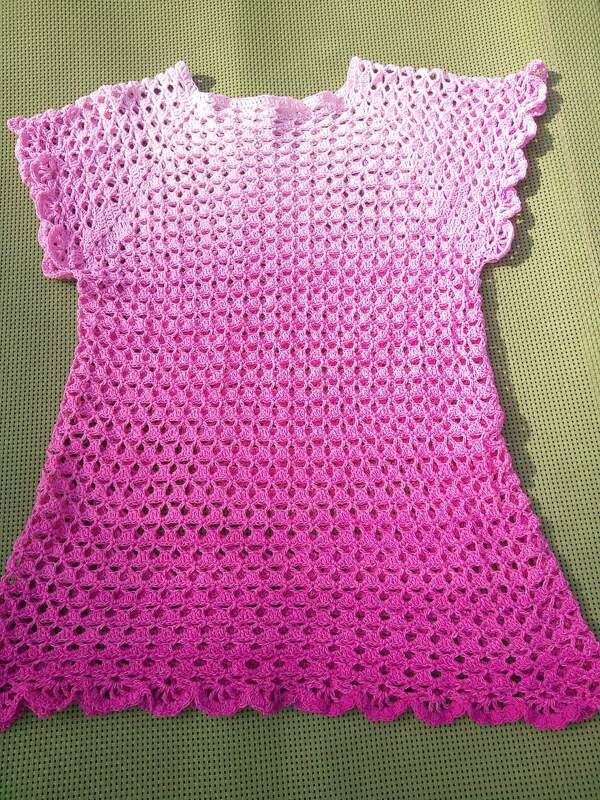Gehäkeltes kurzärmliges Damenshirt in Rosa- und Pinktönen mit Muschelmuster liegt flach auf grünem Untergrund.