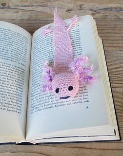 Rosa gehäkeltes Axolotl-Lesezeichen steckt in einem aufgeschlagenen Buch auf einem Holztisch.