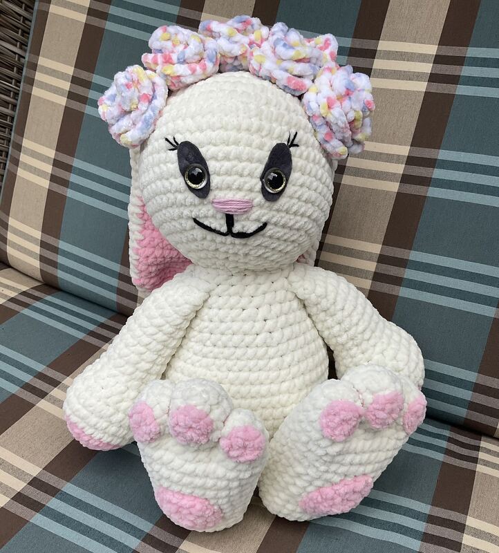Weißer, gehäkelter Amigurumi-Hase mit rosa Ohren und Pfoten, trägt einen pastellfarbenen Blütenkranz und sitzt auf einem karierten Kissen.