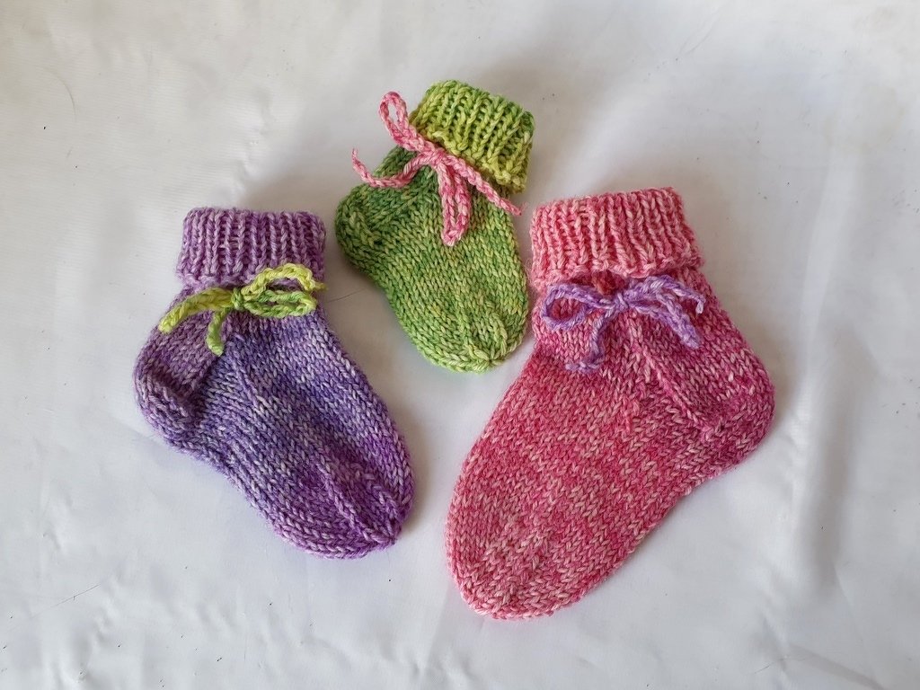 Drei gestrickte Babysocken (lila, grün, rosa) mit Umschlagbündchen und Bändchen auf weißem Hintergrund