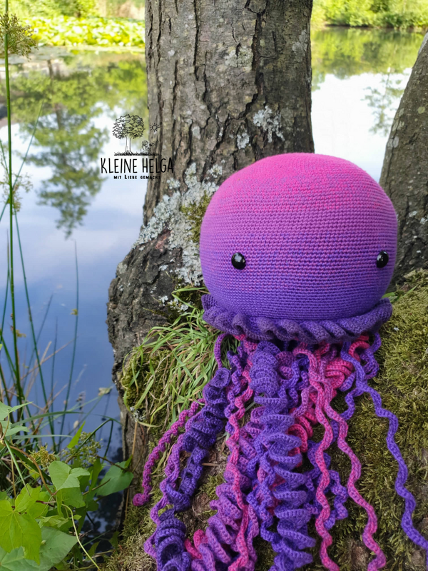 Gehäkelte Amigurumi-Qualle in Lila und Pink mit schwarzen Sicherheitsaugen auf moosigem Baumstamm am Seeufer