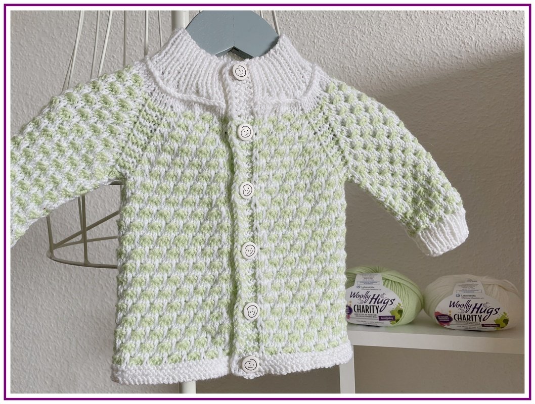 Gestrickte Babyjacke in Weiß und Hellgrün mit durchgehender Knopfleiste und gerippten Bündchen, aufgehängt an einem Kleiderbügel.