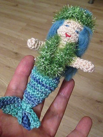 Gehäkelte Amigurumi-Meerjungfrau mit blauen Haaren und blau-grüner Flosse, gehalten in einer Hand.