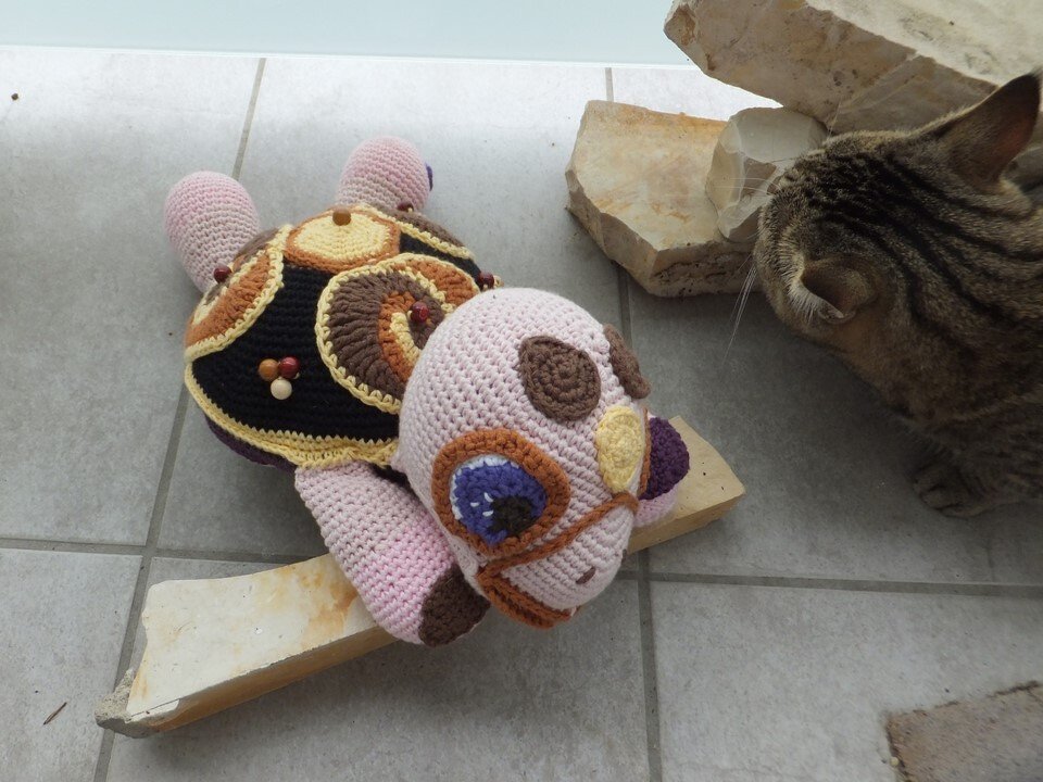 Gehäkelte Amigurumi‑Schildkröte mit gemustertem Panzer und Holzperlen, liegt auf dem Boden neben einer Katze.