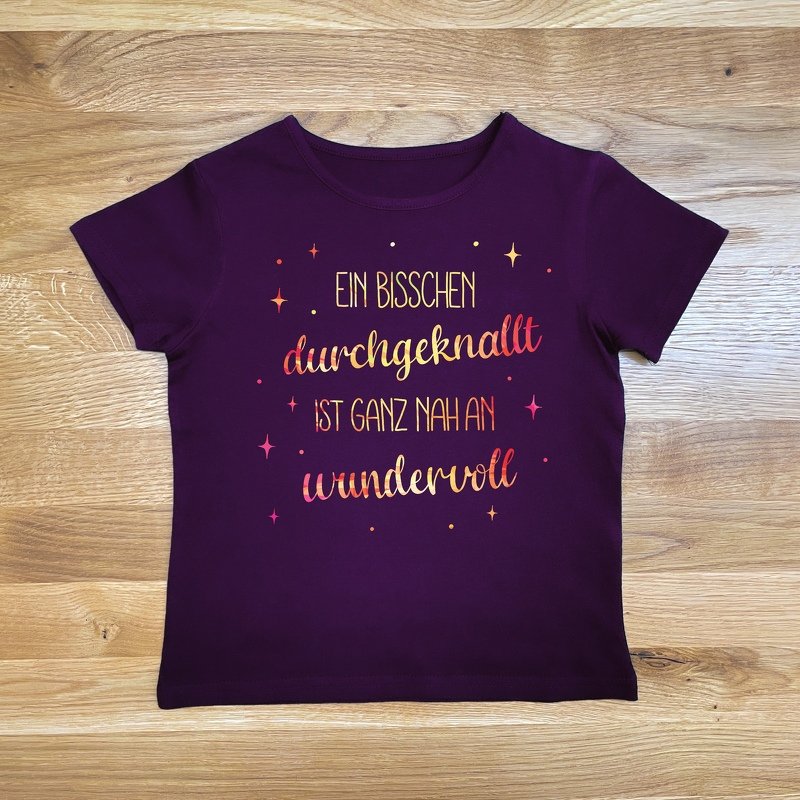 Dunkelviolettes T‑Shirt mit mehrfarbigem Lettering: „Ein bisschen durchgeknallt ist ganz nah an wundervoll“ und Sternakzenten