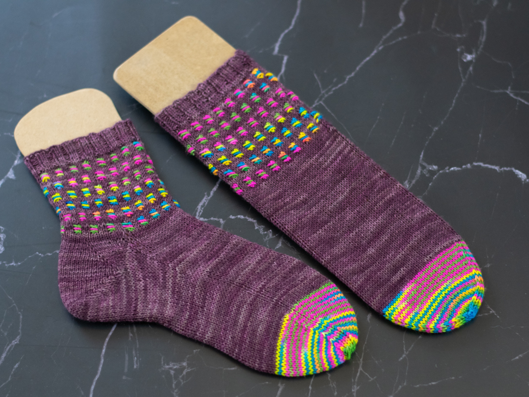 Zwei unterschiedlich lange violette Stricksocken mit gestreiften Zehen und mehrfarbigem Schaftmuster