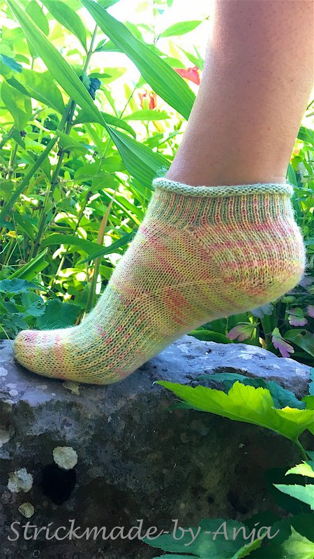 Kurz gestrickte Socke in Grün‑Gelb‑Rosa an einem Fuß auf einem Stein, umgeben von Pflanzen