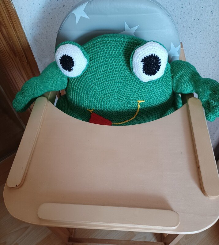 Grünes, gehäkeltes Froschkissen mit großen Augen im Kinderhochstuhl.