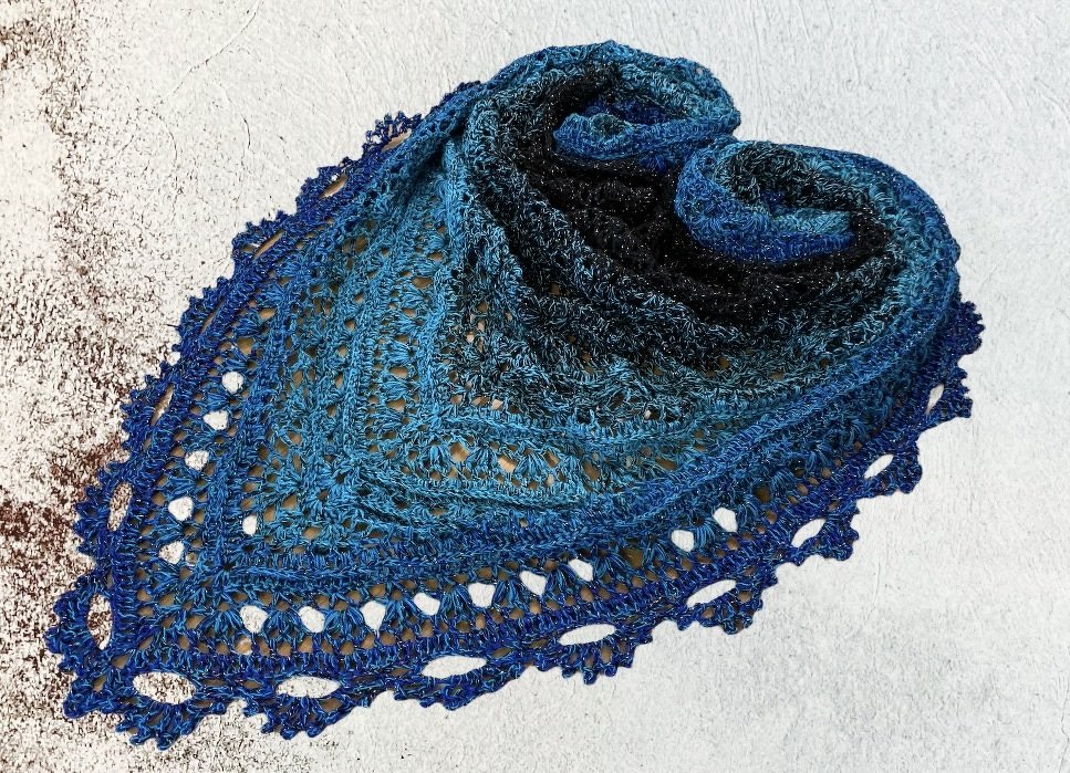 Folded blue ombre crocheted triangle shawl with openwork stitches and scalloped lace edge on a light textured surface.