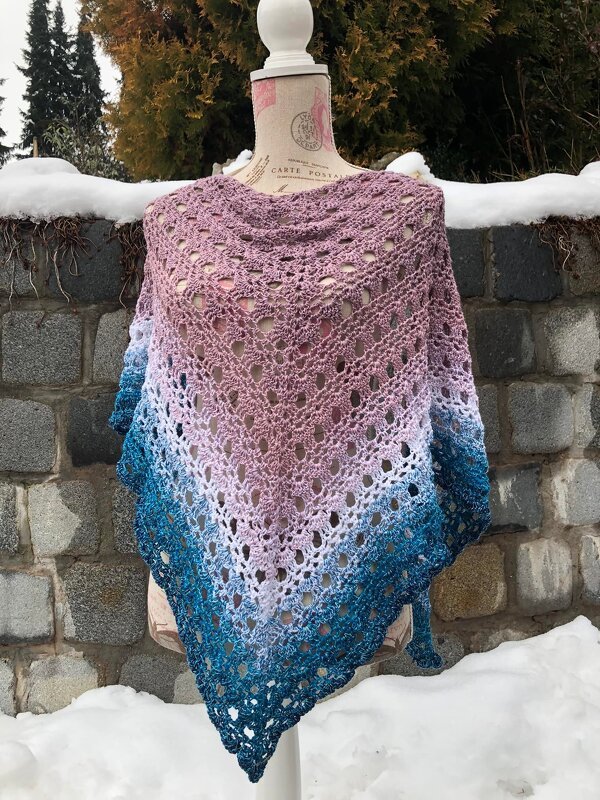 Gehäkeltes Dreieckstuch auf einer Schneiderpuppe mit Farbverlauf von Rosa über Weiß zu Blau, vor einer Steinmauer im Schnee.