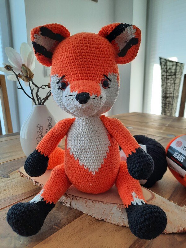 Gehäkelter Amigurumi-Fuchs in Orange, Weiß und Schwarz mit großen Ohren und gestickten Schnurrhaaren, sitzend auf einer Holzscheibe