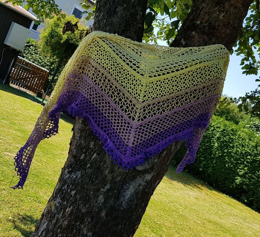 Gehäkeltes Dreieckstuch in Gelb–Lila mit Spitzenmuster, an einem Baumstamm drapiert