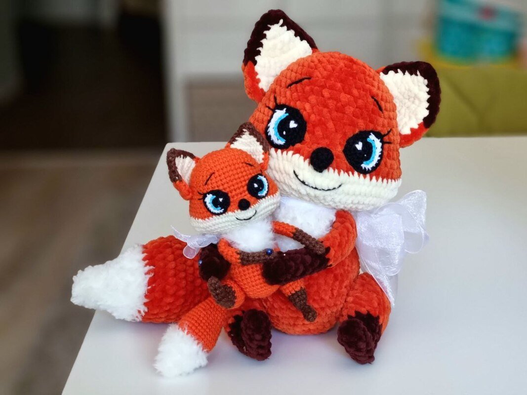 Two crocheted fox amigurumi, larger fox hugging smaller fox, orange and white with blue eyes and white ribbon, seated on a white surface.