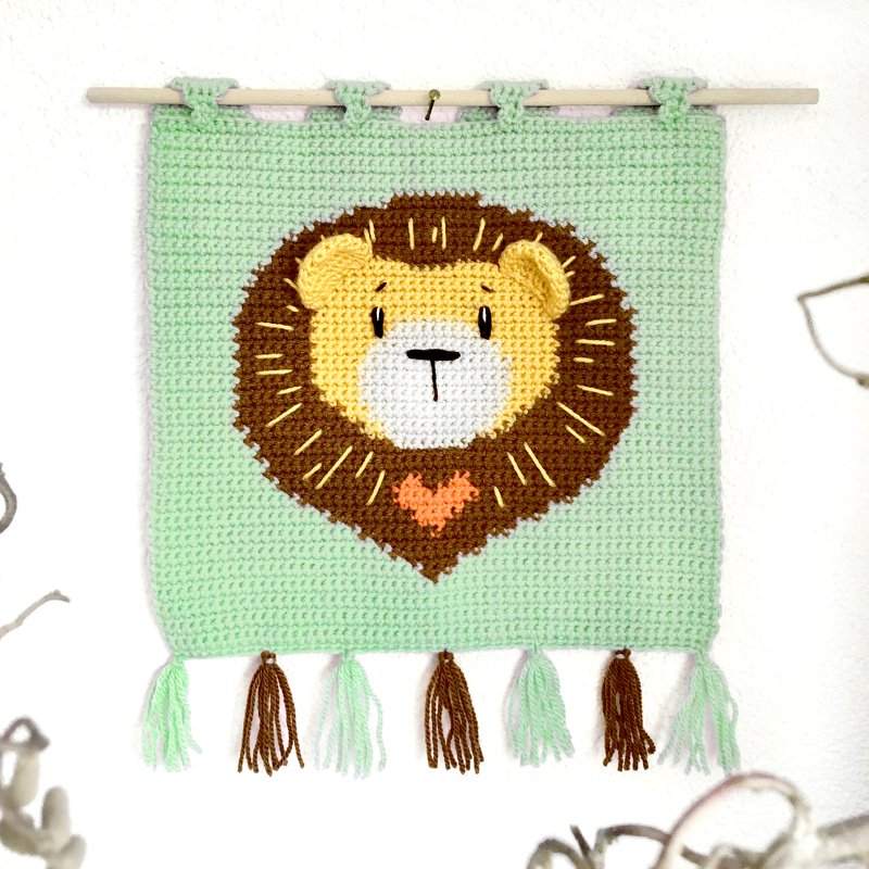 Handgehäkelter Wandbehang in Mintgrün mit gelbem Löwenkopf, brauner Mähne, orangefarbenem Herz und Quasten, an einem Holzstab aufgehängt.