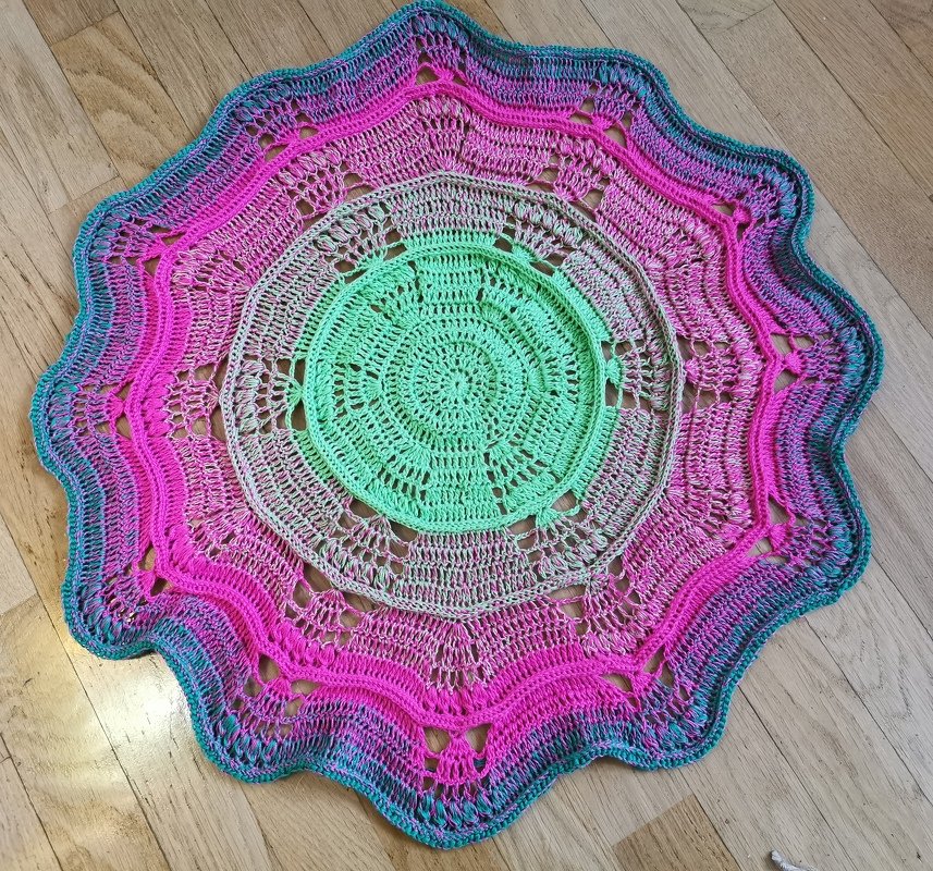 Rundes gehäkeltes Mandala-Deckchen mit Farbverlauf von Grün über Pink zu Türkis auf Holzfußboden