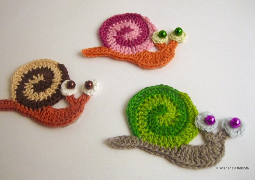 Drei gehäkelte Schneckenapplikationen mit bunten Spiralhäuschen und Perlenaugen auf hellem Untergrund
