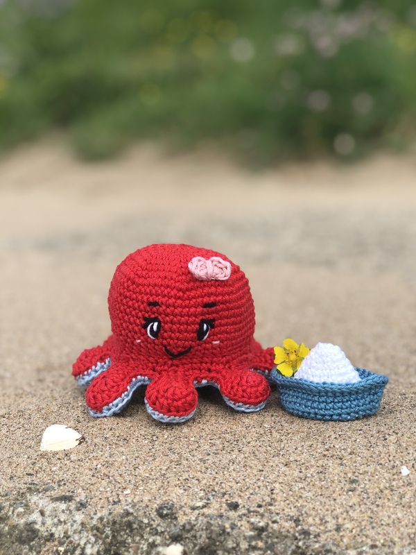 Gehäkelter roter Oktopus mit rosa Schleife, blauer Schale, gelber Blüte und Muschel auf sandigem Boden