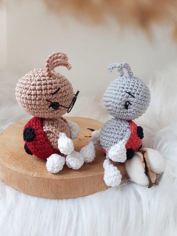 Two crocheted ladybug amigurumi with red spotted shells and white limbs; one wears small round glasses, displayed on a wooden slice.