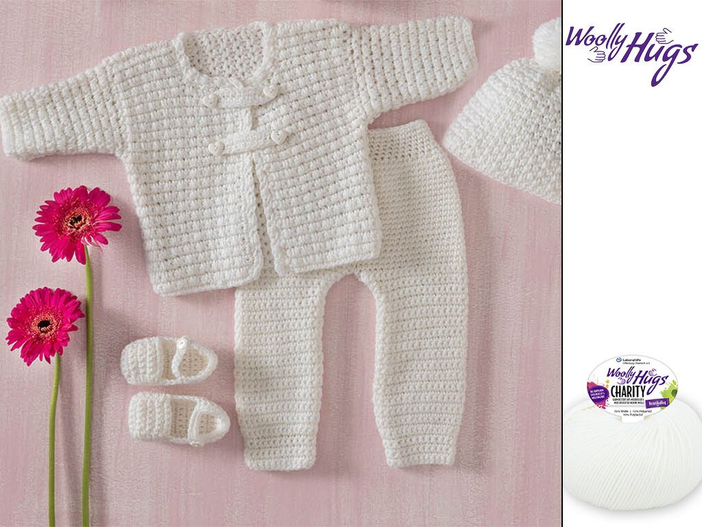 Weißes gehäkeltes Baby-Set mit Jacke, Hose, Mütze und Schühchen auf rosa Untergrund neben zwei pinken Blumen
