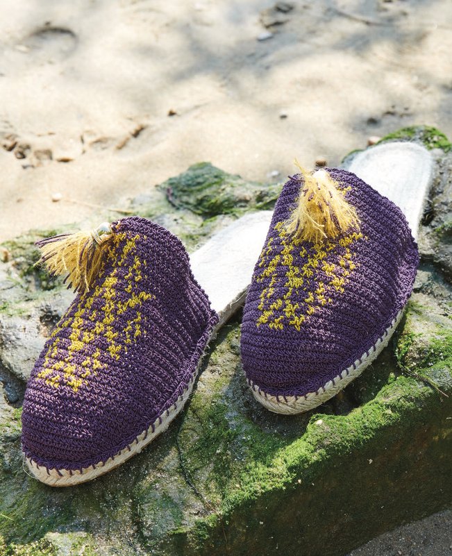 Paar gehäkelter violetter Pantoletten mit gelbem Muster und Quaste auf steinigem Untergrund