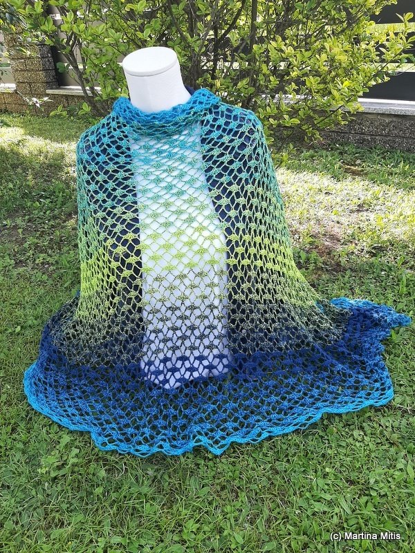 Crocheted triangular shawl in blue, turquoise, and lime green gradient with a lacy mesh stitch, displayed on a mannequin outdoors.