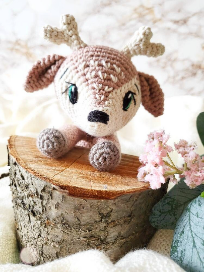 Gehäkeltes Rehkitz-Amigurumi mit Geweih-Ansätzen und grünen Augen auf Holzscheit neben rosa Blüten.