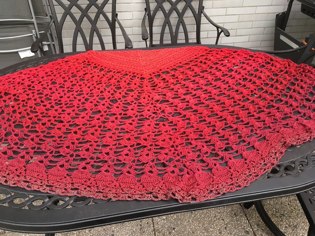 Rotes gehäkeltes Halbrundtuch mit feinem Lochmuster, ausgebreitet auf einem dunklen Gartentisch.