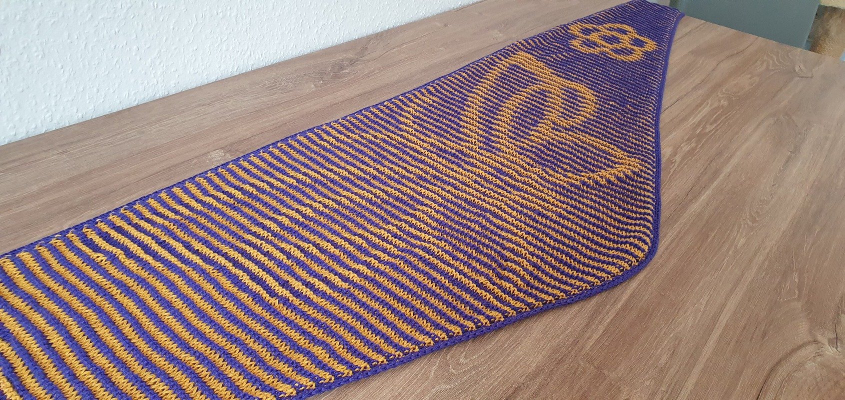 Triangular knitted shawl laid flat with narrow purple and mustard-yellow stripes and a yellow motif at the narrow end