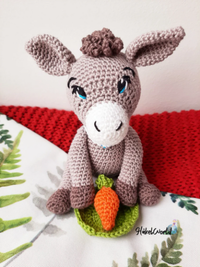Gehäkelter Amigurumi-Esel sitzt und hält eine Karotte auf grünem Blatt
