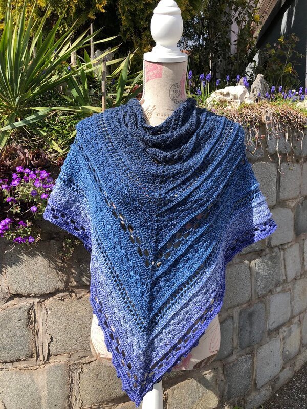 Gehäkeltes Ombré-Dreieckstuch in Blau- und Violetttönen auf einer Schneiderpuppe vor einer Steinmauer und Frühlingsblumen.