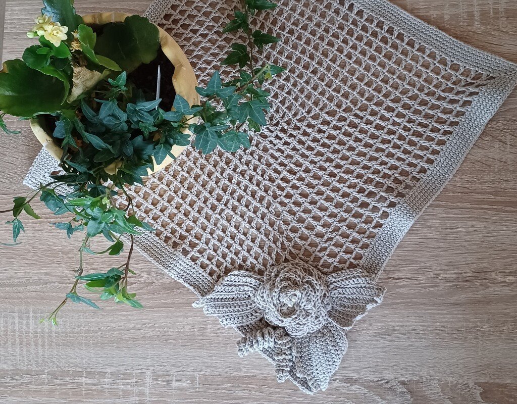 Hellbeiges, gehäkeltes Deckchen mit Gittermuster und gehäkelter Rose mit Blättern neben einer Topfpflanze auf hellem Holztisch.