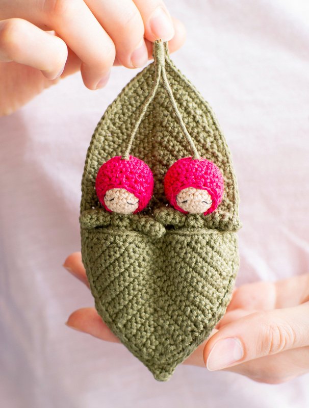 Close-up of hands holding a green crocheted pea pod containing two small amigurumi dolls with pink caps
