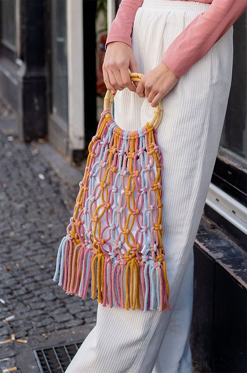 Person hält Makramee-Tasche in Rosa, Hellblau und Gelb mit runden Holzgriffen und Fransen