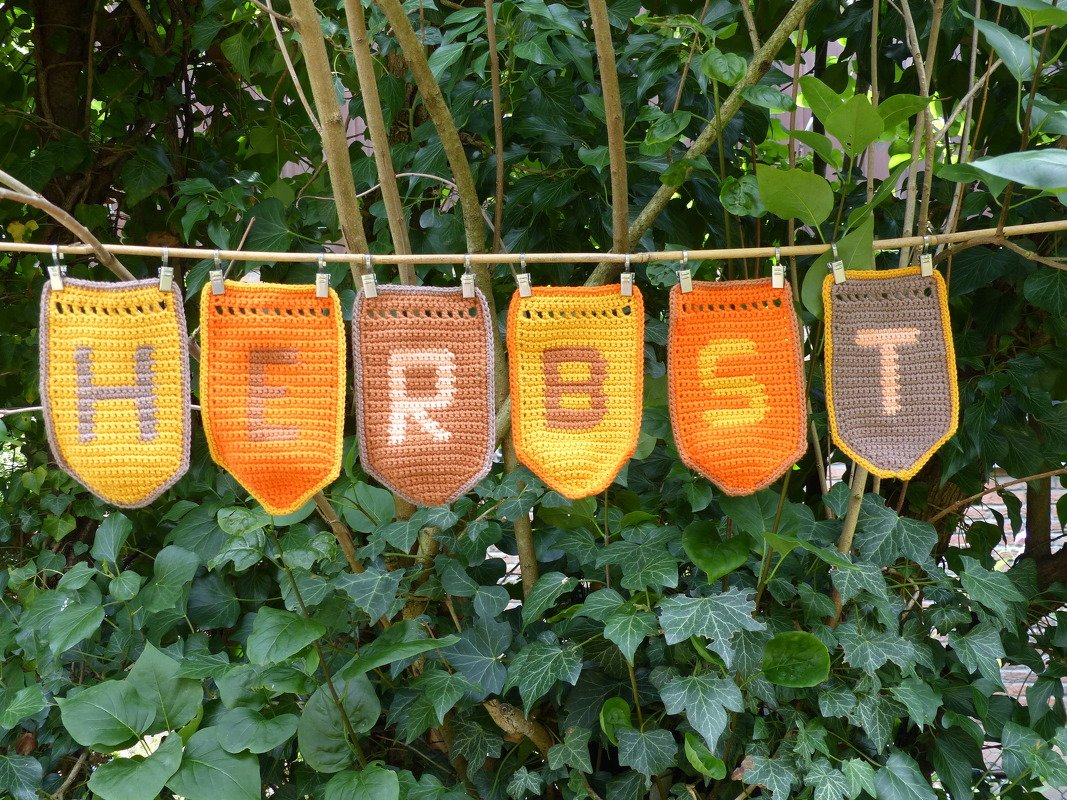 Gehäkelte Wimpelkette mit sechs Wimpeln, die das Wort HERBST in warmen Gelb‑, Orange‑ und Brauntönen zeigen, aufgehängt an einer Schnur vor grünem Blattwerk