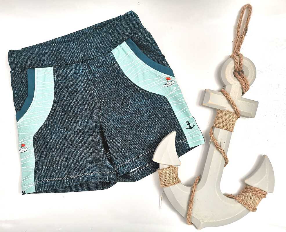 Dunkelblaue Kinder-Shorts mit hellblauen, maritim gemusterten Seitenpartien, daneben ein weißer Deko-Anker.