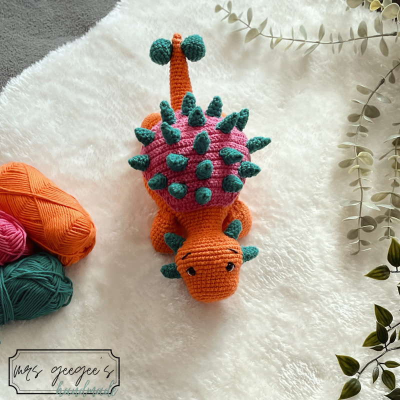 Gehäkelter Amigurumi-Dinosaurier mit rosa Panzer und türkisen Stacheln, orange Körper, auf weißem Plüsch liegend neben Garnknäueln
