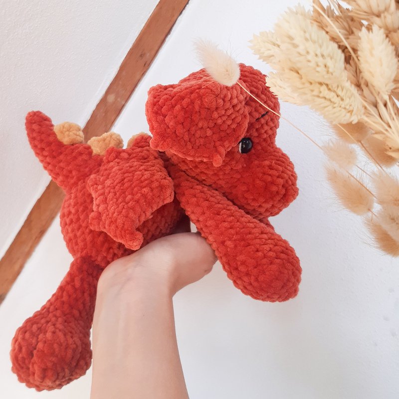 Hand hält einen rostroten, gehäkelten Amigurumi‑Drachen mit Flügeln, Rückenstacheln und Horn.