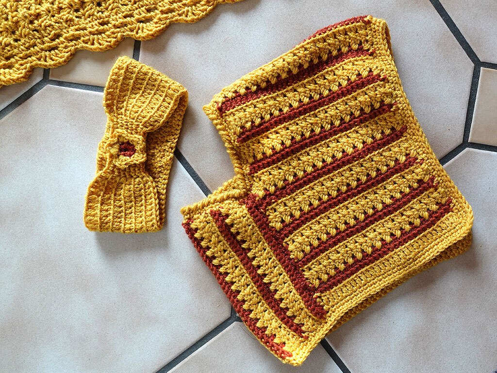 Gehäkeltes Baby-Stirnband mit Blüte und eine weiche, gestreifte Babydecke in Senfgelb und Rostrot auf Fliesenboden.