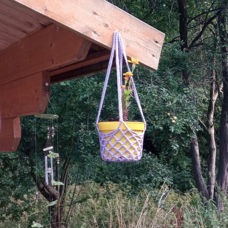 Lila gehäkelte Blumenampel mit Rautenmuster hält einen gelben Blumentopf mit blühender Pflanze, hängend an einem Holzbalken im Garten.