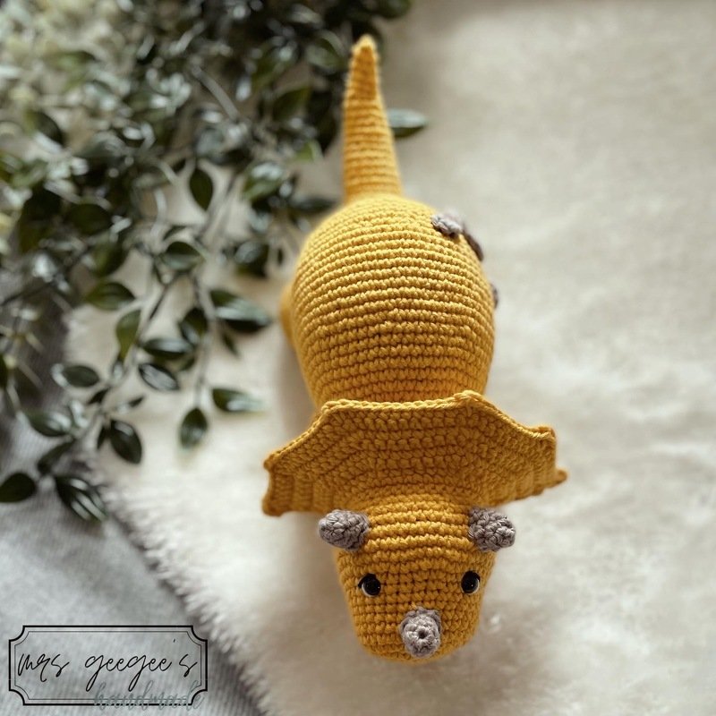 Mustard yellow crocheted triceratops amigurumi with a head frill and grey horns, top-down view