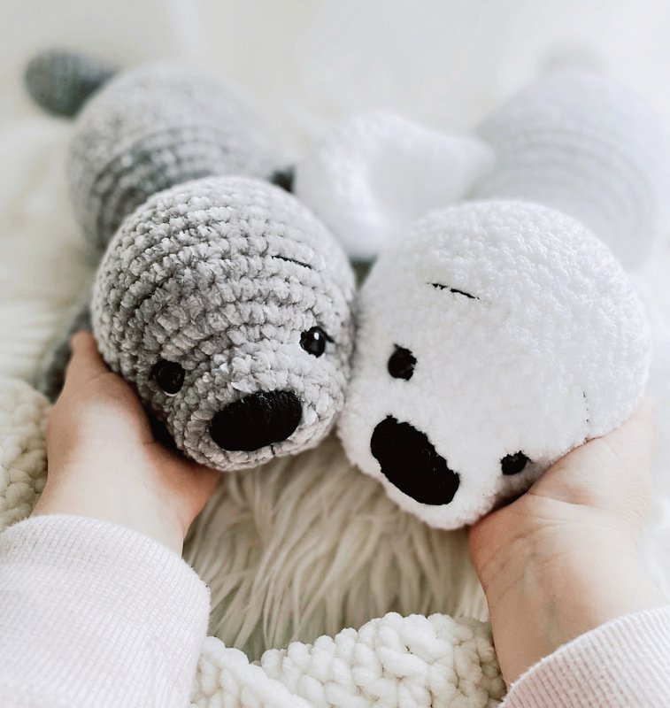Hände halten zwei flauschige gehäkelte Robben-Amigurumi, eine grau und eine weiß, mit schwarzen Augen und Nasen.