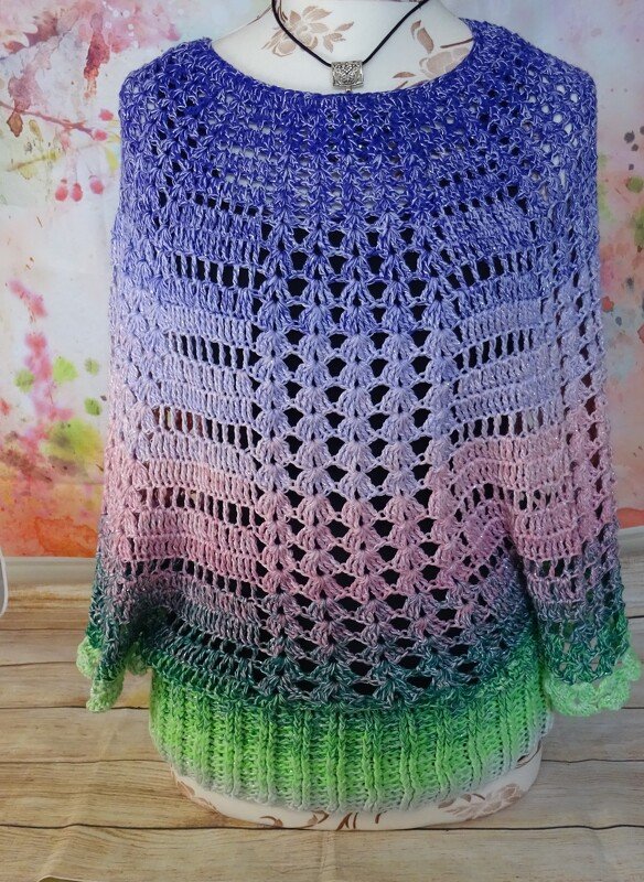 Vorderansicht eines gehäkelten Ponchos mit lila-rosa-grünem Farbverlauf, Lochmuster und geripptem Saum auf einer Schneiderpuppe