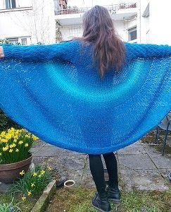 Rückansicht einer Person mit ausgebreiteten Armen, trägt eine halbkreisförmige gehäkelte blaue Jacke mit Farbverlauf