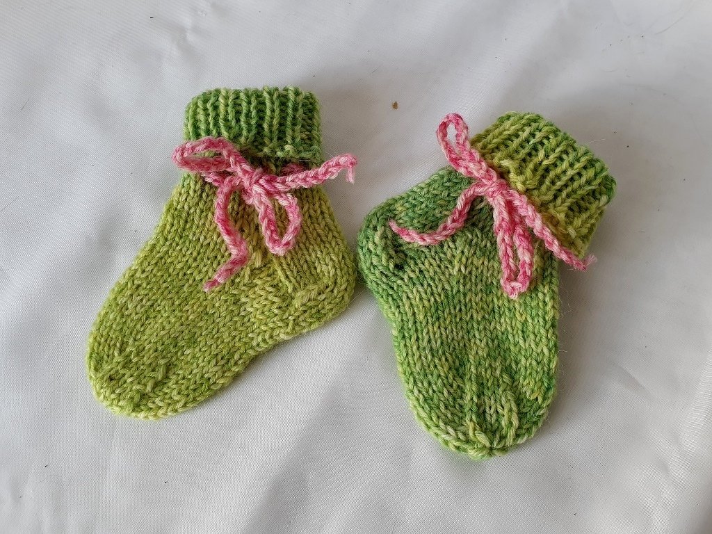 Zwei grüne, gestrickte Babysocken mit geripptem Bündchen und rosa Bindeband auf hellem Untergrund