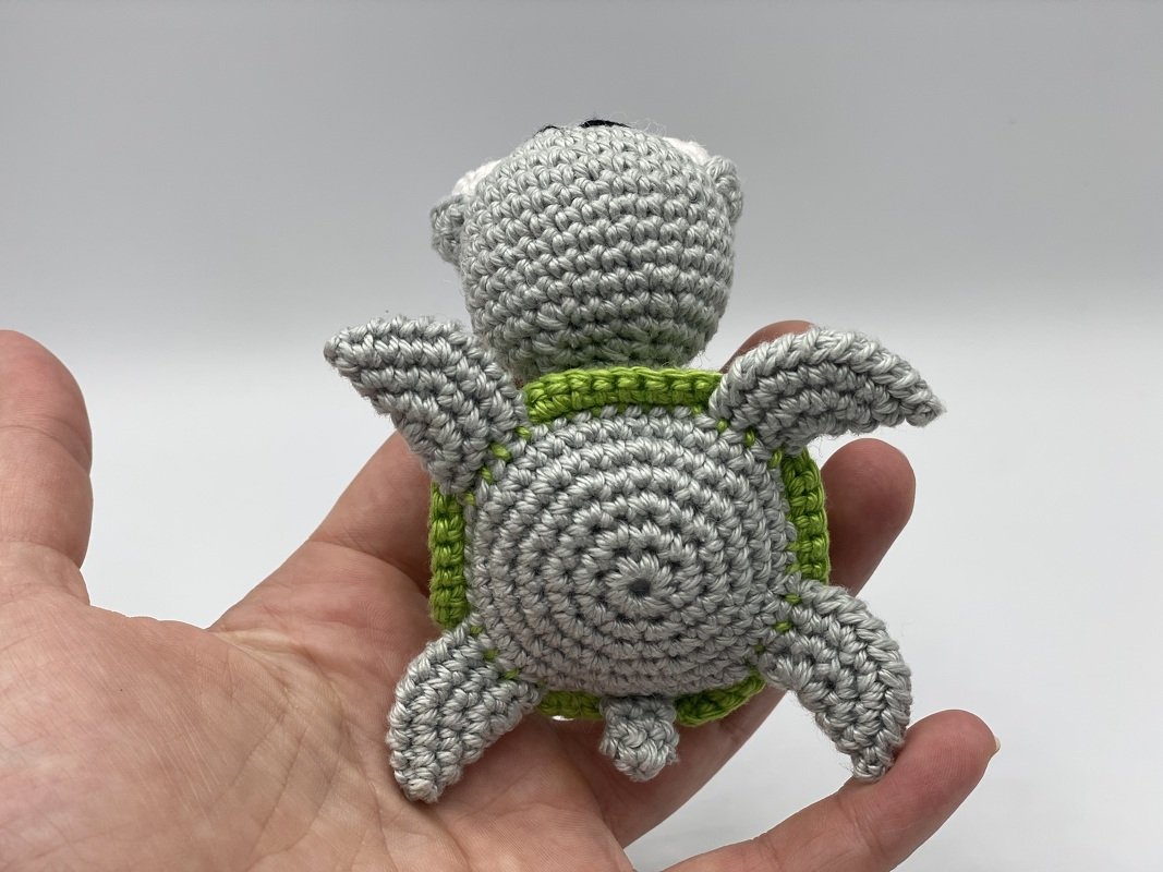 Hand holding a small crocheted turtle with a grey body and green shell, viewed from the back.