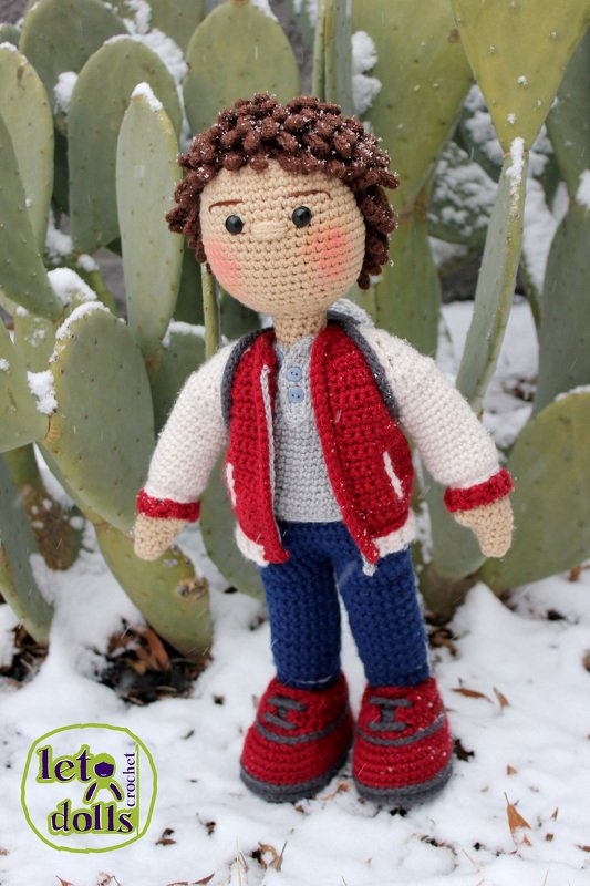 crocheted amigurumi boy doll with curly brown hair wearing a red jacket standing on snow by a cactus