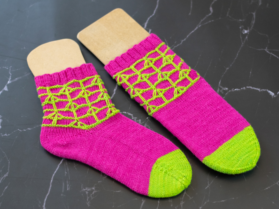 Zwei gestrickte Socken in Pink mit neongrünen Zehen und gitterartigem Lochmuster auf dunklem marmoriertem Untergrund.