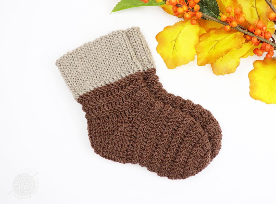 Gehäkelte braun-beige Babysocke auf weißem Hintergrund neben herbstlicher Dekoration