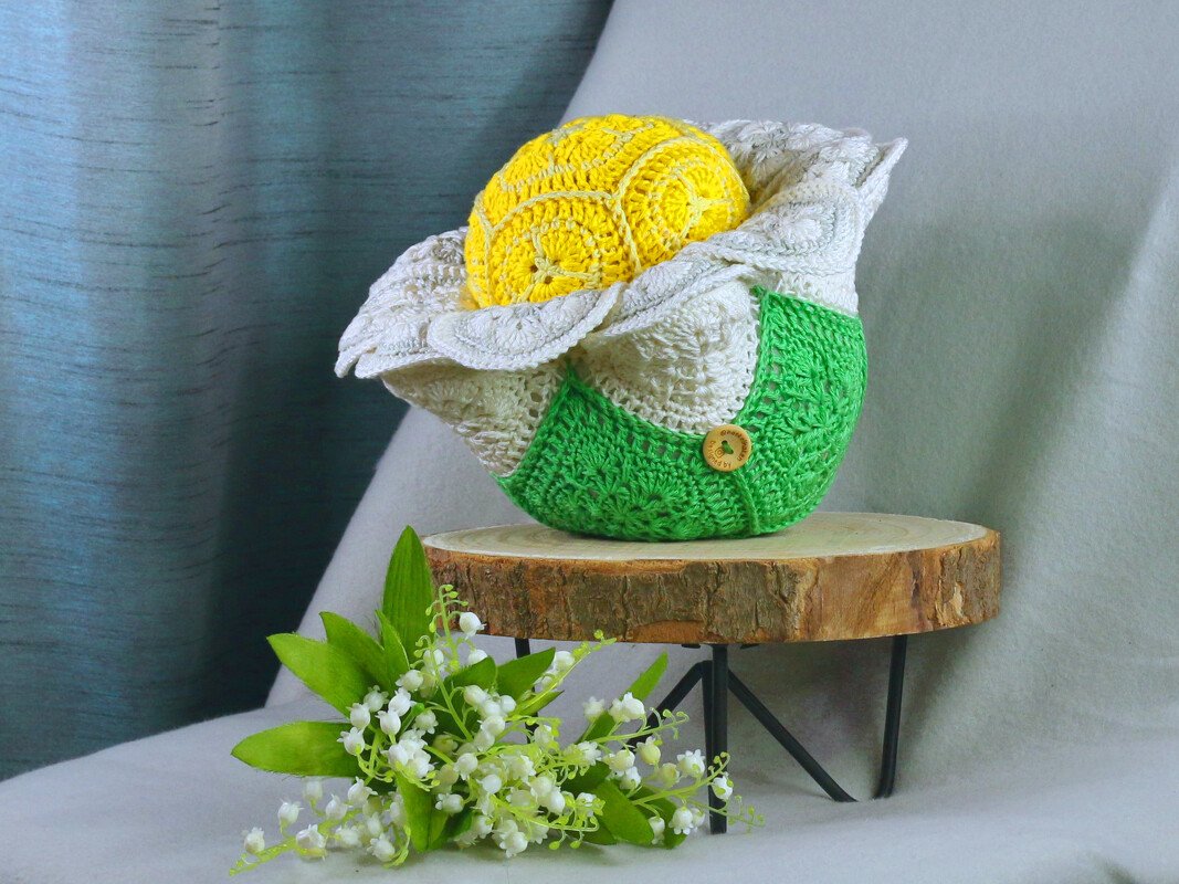 Detailreich gehäkelter Blumenkorb in Grün, Weiß und Gelb mit gelbem Innenkern und Holzknopf auf Holzpodest