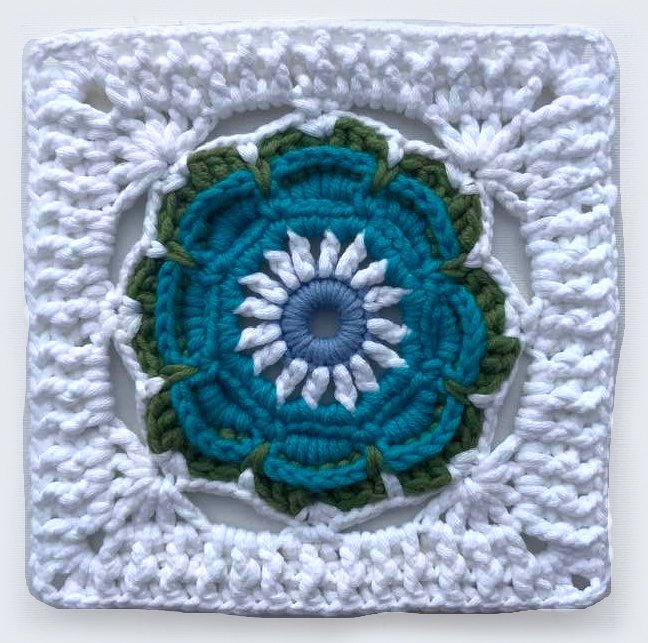 Nahaufnahme eines quadratischen Häkelmotivs mit Blumenmuster in Blau, Türkis, Weiß und Grün, umgeben von weißem Rand.
